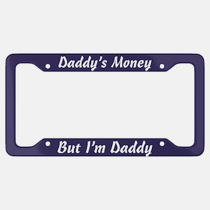 May include: A dark blue license plate frame with white text that reads "Daddy's Money" at the top and "But I'm Daddy" at the bottom. The frame has rounded corners and small white dots.