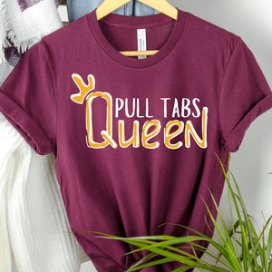 May include: Burgundy t-shirt with white text that says "Pull Tabs Queen" with a gold crown above the text.