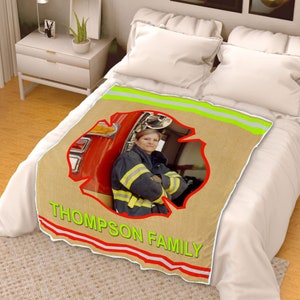 Pode incluir: Uma manta de arremesso bege com um design de hidrante de incêndio vermelho. O design apresenta uma foto de um bombeiro em pé na frente de um caminhão de bombeiros. A manta tem o texto "THOMPSON FAMILY" em letras vermelhas e brancas.
