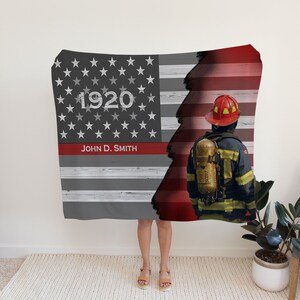 Pode incluir: Uma manta de bandeira americana cinza e vermelha com um bombeiro em pé na frente dela. A bandeira está rasgada no meio, revelando as listras vermelhas da bandeira. O bombeiro está usando um capacete e um uniforme amarelo e preto. A manta tem o texto "1920" e "John D. Smith" nela.