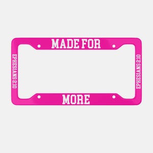 以下が含まれることがあります： 鮮やかなピンク色のナンバープレートフレーム。白い文字で「MADE FOR MORE」と書かれています。テキスト「EPHESIANS 2:10」が側面に縦書きで印刷されています。フレームにはナンバープレート用の4つの取り付け穴があります。