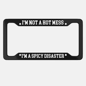 Peut inclure: Cadre de plaque d'immatriculation noir avec texte blanc. Le texte supérieur indique "I'M NOT A HOT MESS" et le texte inférieur "I'M A SPICY DISASTER". Le cadre est rectangulaire.