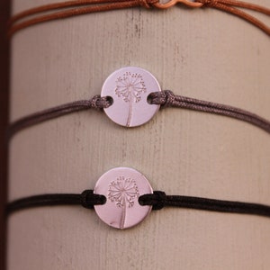 Könnte beinhalten: Drei Armbänder mit runden, silberfarbenen Anhängern mit einem Pusteblumen-Design. Die Armbänder haben verstellbare Kordelbänder in Braun, Grau und Schwarz. Die Armbänder werden auf einer hellen zylindrischen Oberfläche präsentiert.