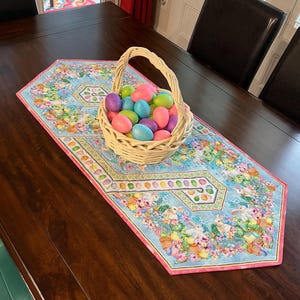 May include: A light blue Easter table runner with a pink border, featuring a floral and bunny design. A woven basket filled with colorful, glittery Easter eggs sits on the runner. The table is made of dark wood.