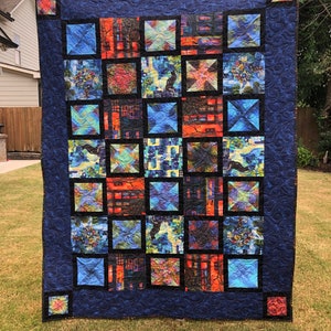 May include: A handmade quilt with a dark blue border and a grid of colorful squares. The squares feature abstract designs in shades of blue, orange, and green. The quilt is displayed outdoors on a grassy lawn.