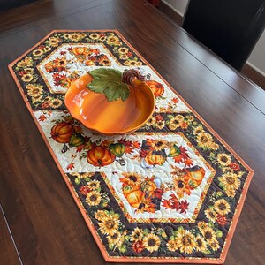 May include: A quilted table runner with a fall theme, featuring pumpkins, sunflowers, and autumn leaves. The runner has a white background with a repeating pattern of pumpkins and leaves. A ceramic pumpkin-shaped bowl sits on top.