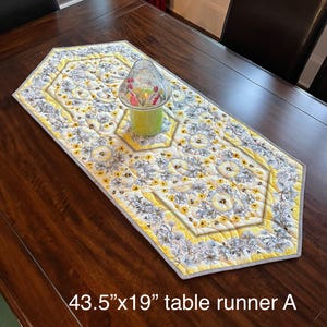 May include: A yellow, gray, and white floral quilted table runner, measuring 43.5" x 19", is displayed on a dark wood table. The runner features a repeating floral pattern and a geometric design. A candle holder with a floral design sits on the runner.