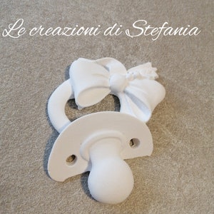Puede incluir: Un chupete de cerámica blanco para bebé con un gran lazo blanco en la parte superior. El chupete es un regalo popular para baby shower.