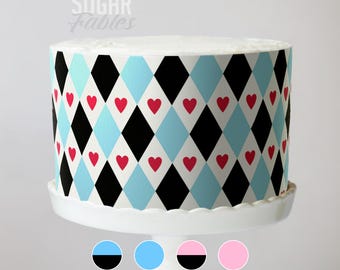 May include: A round cake with a white top and a patterned exterior. The pattern features black and light blue diamonds with red heart accents. The cake sits on a white cake stand. The text "Sugar Fables" is visible.