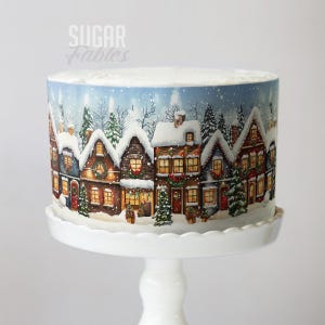 May include: A round cake with a winter village scene printed on the side, set on a white cake stand. The village features snow-covered houses with lit windows, Christmas trees, and a snowy sky. The cake is topped with white frosting.