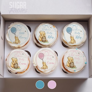 May include: A box containing six cupcakes, each topped with white icing and edible images. The images depict Winnie the Pooh with balloons and the text "Welcome Baby Riley", "Welcome Baby Emma", and "What will Baby Bee?" The cupcakes are in a white box.