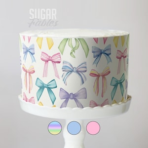 May include: A round cake with a white base, decorated with watercolour bows in various colours, including blue, pink, yellow, and green. The cake sits on a white cake stand. The text "Sugar Fables" is at the top.