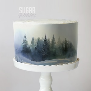 May include: A round cake with a winter forest scene painted on the side, displayed on a white cake stand. The cake features a snowy landscape with evergreen trees in shades of blue and green. The text "Sugar Fables" is visible on the top left.