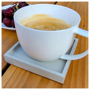 May include: A white ceramic coffee mug with a light brown coffee drink inside, sitting on a gray square concrete coaster on a wooden surface.