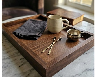 May include: A dark brown wooden serving tray with a rectangular shape. The tray holds a gray linen napkin, a cream-colored mug with a gold rim, a small silver cup, and two gold-colored stir sticks. A book is visible in the background.