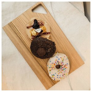 Może przedstawiać: Trzy pączki na drewnianej desce do krojenia. Jeden pączek jest polany bekonem, kompotem z borówek i pianką. Inny pączek jest czekoladowy z czekoladowym nadzieniem. Trzeci pączek jest biały z kolorowymi posypkami.