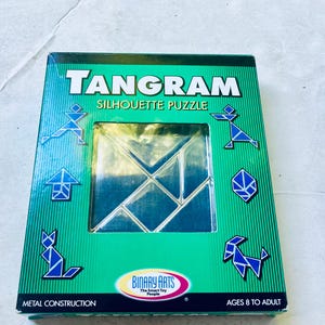 May include: A green rectangular box for a Tangram Silhouette Puzzle by Binary Arts. The box features a central clear window displaying metal puzzle pieces, with blue geometric illustrations of a cat, dog, and human figures on the front.