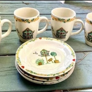 May include: A set of four cream-colored ceramic mugs and a stack of matching plates. Each mug and plate features a birdhouse design with green, red, and blue accents. The mugs have a handle and a decorative border.