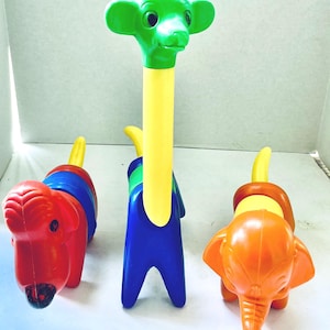 May include: Three colorful plastic animal toys: a red dog, a giraffe, and an orange elephant. The giraffe has a green head and yellow neck. The dog is red with blue and red rings. The elephant is orange with a yellow tail.