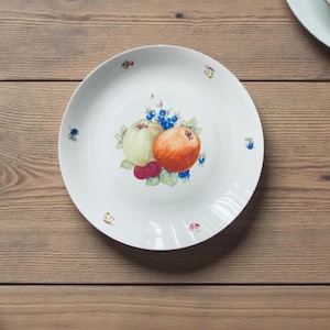 Puede incluir: Plato de cerámica blanca con un diseño de frutas pintado a mano. Presenta una manzana verde y naranja, bayas rojas y arándanos azules. El plato tiene un diseño simple y elegante, ideal para servir alimentos.