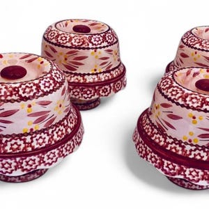 以下が含まれることがあります： 花柄の装飾的なセラミッククローシュ4つ。クローシュは白地にマルーンの花柄とマルーンのベース。クローシュの上部には小さなマルーンのノブがあり、白い背景にあります。