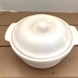 Puede incluir: Una fuente de cerámica blanca con tapa. La fuente tiene dos asas y un pomo en la tapa. La fuente está sobre una superficie de madera marrón.