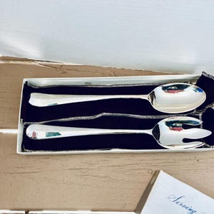 May include: A set of two silver serving utensils, a spoon and a fork, presented in a white box with a navy blue velvet lining. The utensils are highly polished and reflect the surrounding environment.