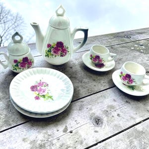 May include: A vintage tea set featuring a white teapot, sugar bowl, two teacups with saucers, and two plates. Each piece is decorated with a floral design in shades of purple and pink. The set is arranged on a weathered wooden surface.