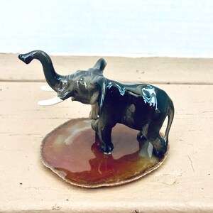 May include: A glossy, dark gray ceramic elephant figurine with white tusks. The elephant stands on a brown, oval-shaped base. The figurine has a reflective surface, and the trunk is raised. The overall design is detailed and elegant.