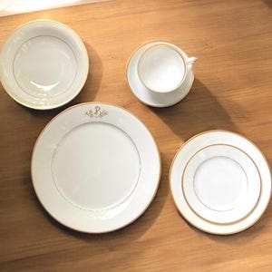 Peut inclure: Un service de table en porcelaine blanche avec des bordures dorées. L'ensemble comprend un bol, une tasse et une soucoupe, une assiette avec un monogramme doré et une petite assiette. Les articles sont disposés sur une surface en bois.