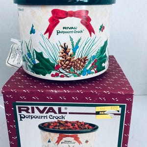 Puede incluir: Un recipiente para popurrí Rival con tapa verde oscuro y cuerpo color crema decorado con un lazo rojo, piñas y bayas de acebo. El recipiente se asienta sobre su caja original color granate, que muestra el nombre y una ilustración.