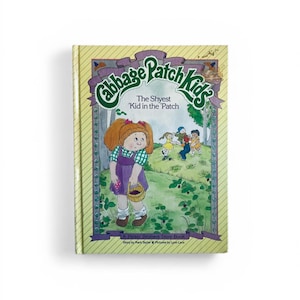 Könnte beinhalten: Ein Vintage-Cabbage-Patch-Kids-Geschichtenbuch mit dem Titel "The Shyest 'Kid in the 'Patch". Das Cover zeigt eine Cartoon-Illustration eines Mädchens in einem lila Kleid und einem Korb, mit anderen Kindern, die im Hintergrund spielen.