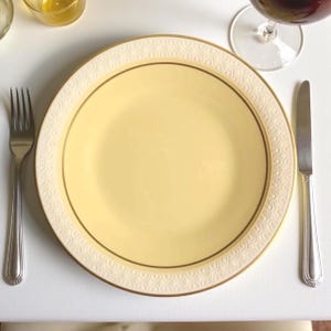 Op de afbeelding: Een gele plaat met een gouden rand en een wit patroon, samen met een vork en een mes. De plaat staat op een wit oppervlak, wat wijst op een formele eetgelegenheid. Een glas rode wijn is zichtbaar op de achtergrond.