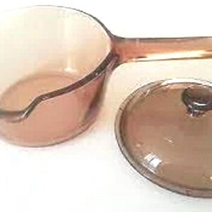 May include: A clear, amber-colored saucepan with a handle and a matching lid. The saucepan has a pouring spout and is made of glass. The lid has a dark brown knob. Kitchenware.