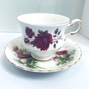 May include: White teacup and saucer set with a floral design. The teacup features a large burgundy rose and smaller buds, with gold trim on the handle and rim. The saucer has a matching design with thistles.