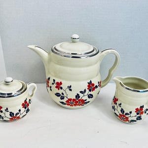 Puede incluir: Juego de té color crema con estampado floral rojo. El juego incluye una tetera, un azucarero y una lechera, cada uno con ribete plateado. La tetera tiene tapa y boquilla curva. El azucarero tiene tapa y dos asas. La lechera tiene asa.