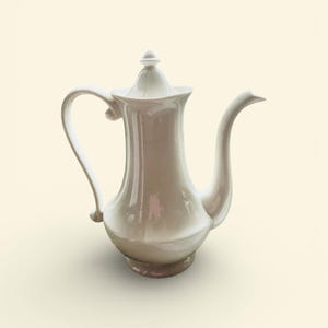 May include: A white ceramic coffee pot with a curved handle and spout. The pot has a lid with a small knob and sits on a round base. The design is elegant and classic, suitable for serving coffee or tea.