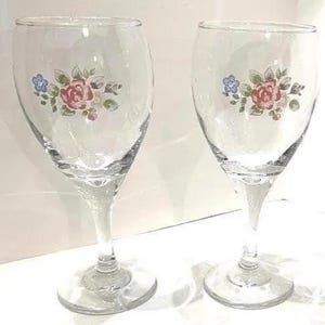 May include: Two clear wine glasses with floral patterns. Each glass has a design of pink roses, blue flowers, and green leaves. The glasses have a traditional stem and a rounded bowl, ideal for serving wine.