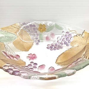 May include: A clear glass serving dish with a decorative grape and leaf design. The dish features clusters of purple grapes, gold and green leaves, and pink flowers. The rim of the dish has a scalloped edge.