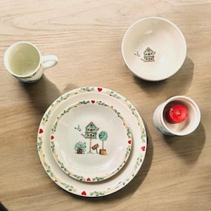 May include: A collection of ceramic tableware with a birdhouse design. Includes a mug, a bowl, a small candle holder with a red candle, and two plates. The plates and bowl have a green leaf and red heart border.