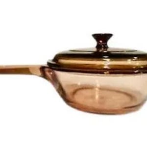 Könnte beinhalten: Ein klarer Glas-Kochtopf mit langem Griff und passendem Deckel. Topf und Deckel haben einen hellen Bernsteinton. Der Deckel hat einen dunkelbraunen Knopf. Der Kochtopf ist für das Kochen auf dem Herd konzipiert.