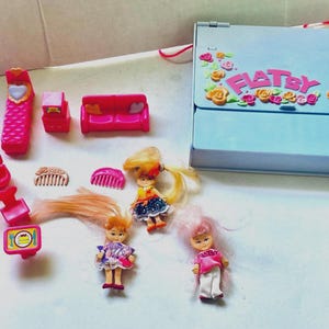 Könnte beinhalten: Eine Sammlung von Miniaturspielzeugen im Vintage-Stil, darunter Puppen, Möbel und Zubehör. Das Set enthält einen hellblauen Tragekoffer mit dem Wort "FLATSY" und floralen Verzierungen. Die Puppen haben bunte Outfits und Frisuren. Die Möbel sind hauptsächlich rosa.