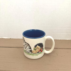 Op de afbeelding: Crèmekleurige keramische mok met een blauwe binnenkant en een witte handgreep. De mok heeft een kattenillustratie aan de buitenkant, met een grijze en witte kat die naast een kitten in een nest zit. Het nest bevat rode bessen en groene bladeren.