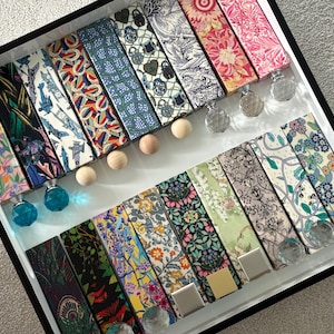 May include: A selection of decorative bottle stoppers with diverse floral and patterned designs. The stoppers feature wooden or crystal accents and are arranged in a black tray. The tray is on a white surface.