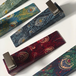 May include: Several rectangular fabric cases with metal clips, each displaying a peacock feather pattern. The cases are in shades of teal, navy, burgundy, and green. The metal clips are silver.