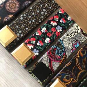 May include: A selection of patterned drawer pulls. The designs include floral, celestial, and peacock feather patterns. Each pull has a gold-coloured square base. The pulls are made of fabric.