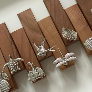 May include: A collection of silver decorative knobs, each fixed to a rectangular wooden block. Designs include a pineapple, turtle, guitar, bird, wings, dragonfly, palm tree, and seashell. The wood has a warm, reddish-brown colour.