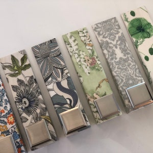 May include: A selection of fabric napkin rings with diverse floral and botanical patterns. Each ring has a rectangular silver-toned metal clasp. The patterns include blue, yellow, green, and grey floral designs.