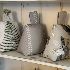 May include: Three fabric doorstops in neutral tones. One is adorned with a green fern print, another with a white leaf pattern, and the third with a floral design in blue and white. Each has a fabric loop handle for easy carrying.