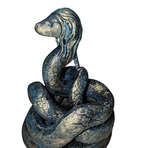 Pode incluir: Uma figura de cobra de cerâmica dourada e azul com uma cabeça detalhada e um corpo enrolado. A cobra está enroscada em forma circular, com a cabeça levantada e olhando para o lado.